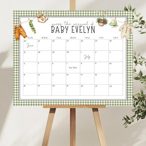 May include: A calendar on a wooden easel with a green gingham border. The calendar grid is for guessing the arrival of Baby Evelyn, with illustrations of baby clothes and greenery. The months of June and July are visible.