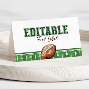 May include: A white, folded card with the words "EDITABLE Food Label" in green and brown. A brown football graphic is on the card. The bottom of the card has a green football field design. The card is on a wooden tray.