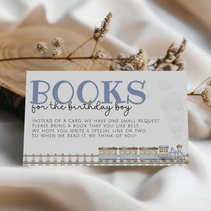 Puede incluir: Tarjeta rectangular con el texto "BOOKS for the birthday boy" en azul. La tarjeta incluye una solicitud para traer un libro y una ilustración de tren en la parte inferior. El fondo es de color neutro.