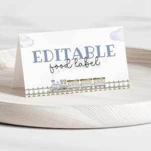 May include: A white, folded card with the text "EDITABLE food label" in blue and black script. A watercolor illustration of a blue train with gold accents and a railroad track is at the bottom. Two blue cloud illustrations are at the top.