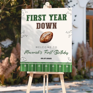 May include: A white sign on a wooden easel with the text "FIRST YEAR DOWN" in green and brown. A football graphic is in the center, with the text "WELCOME TO Maverick's First Birthday" and the date "09.27.2025". The bottom has a green football field design.