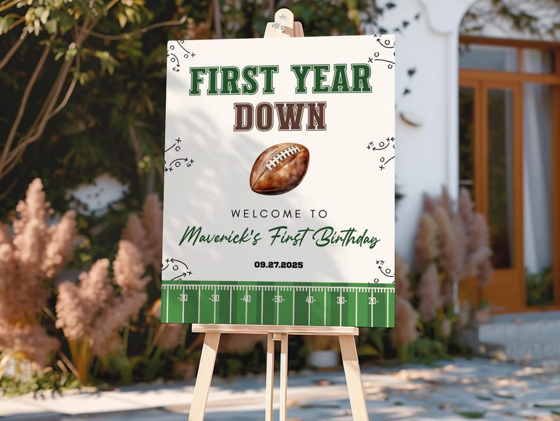 May include: A white sign on a wooden easel with the text "FIRST YEAR DOWN" in green and brown. A football graphic is in the center, with the text "WELCOME TO Maverick's First Birthday" and the date "09.27.2025". The bottom has a green football field design.