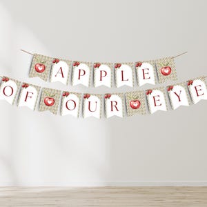Puede incluir: Una guirnalda decorativa con la frase "APPLE OF OUR EYE" en letras rojas. Cada banderín tiene una ilustración de una manzana roja. La guirnalda está ensartada en una fina cuerda marrón.
