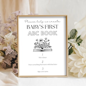 Puede incluir: Un letrero enmarcado en dorado con el texto "BABY'S FIRST ABC BOOK" en negro. El letrero incluye instrucciones para elegir una letra, dibujar algo que comience con esa letra y firmar tu nombre. El fondo presenta flores blancas y moradas.