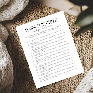 May include: A white paper with the text "PASS THE PRIZE baby edition" is displayed on a woven mat. The text is in black, with a list of instructions below. A white knitted blanket is in the background.