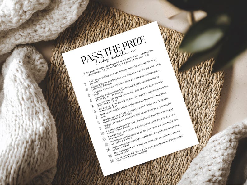 May include: A white paper with the text "PASS THE PRIZE baby edition" is displayed on a woven mat. The text is in black, with a list of instructions below. A white knitted blanket is in the background.