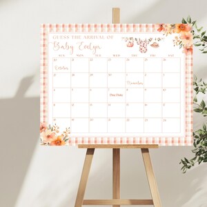 May include: A calendar on a wooden easel with the text "Guess the Arrival of Baby Evelyn." The calendar grid is surrounded by a peach and white gingham border and floral accents. The calendar includes the months of October and November.