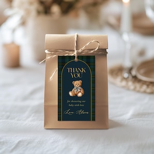 Puede incluir: Una bolsa de regalo de papel marrón atada con hilo, con una etiqueta a cuadros azul marino y verde. La etiqueta dice "THANK YOU" encima de una ilustración de oso de peluche y texto: "for showering our baby with love Love, Alison."