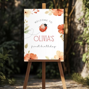 May include: A wooden easel displays a welcome sign for Olivia's first birthday. The sign features a ladybug, floral accents, and the text "Welcome to Olivia's first birthday" in a whimsical font. The sign is surrounded by greenery.