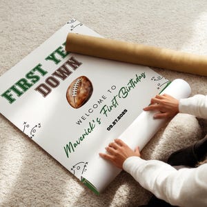 May include: A white poster with green and brown text that reads "FIRST YEAR DOWN" and "WELCOME TO Maverick's First Birthday" with a football graphic. The date "09.27.2025" is also printed on the poster. A rolled-up poster is next to it.