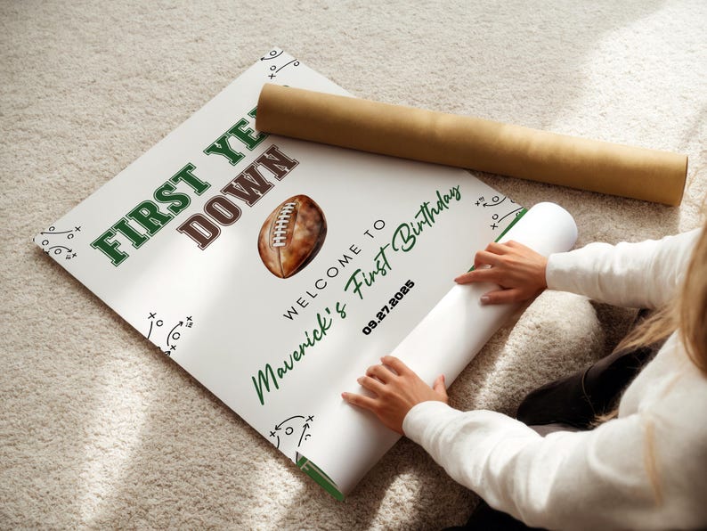 May include: A white poster with green and brown text that reads "FIRST YEAR DOWN" and "WELCOME TO Maverick's First Birthday" with a football graphic. The date "09.27.2025" is also printed on the poster. A rolled-up poster is next to it.