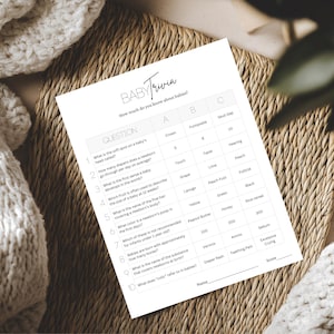 May include: A white baby trivia game sheet with questions and answers, featuring the text "BABY Trivia". The sheet is on a woven mat, with a soft, white blanket in the background. The game is about babies.