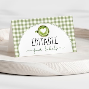 May include: A folded card with a green gingham pattern and the words "EDITABLE food labels" in black and green script. A watercolor image of a green drink with a heart design is on the card. The card is on a light wooden tray.