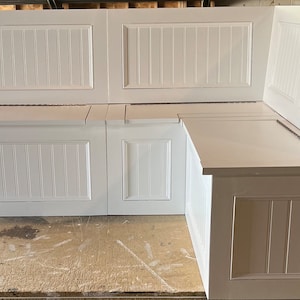 May include: White corner bench with vertical paneling on the sides and back. The bench has a hinged lid for storage and is designed to fit in a corner. The bench is made of wood and painted white.