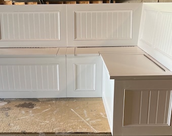 May include: White corner bench with vertical paneling on the sides and back. The bench has a hinged lid for storage and is designed to fit in a corner. The bench is made of wood and painted white.
