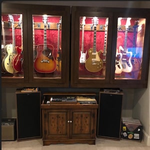 May include: A wooden cabinet with glass doors displays a collection of electric and acoustic guitars. The cabinet has a red velvet interior and is lit from within. The cabinet is flanked by two large black speakers and a smaller amplifier. The cabinet is sitting on a wooden stand with two doors.