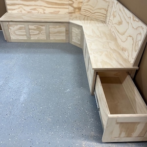 May include: A custom-built, L-shaped wooden bench with a natural finish. The bench features storage compartments with cabinet doors and a drawer that is partially open. The wood grain is visible throughout the design.