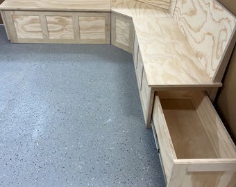 May include: A custom-built, L-shaped wooden bench with a natural finish. The bench features storage compartments with cabinet doors and a drawer that is partially open. The wood grain is visible throughout the design.