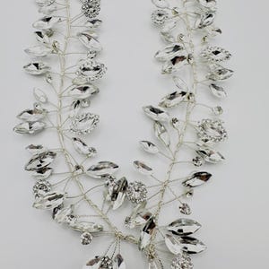 May include: A silver-toned bridal headpiece featuring a delicate vine design. The headpiece is adorned with clear, sparkling crystals in various shapes, including oval and round, and small floral accents. The design is symmetrical and elegant, perfect for weddings or special occasions.
