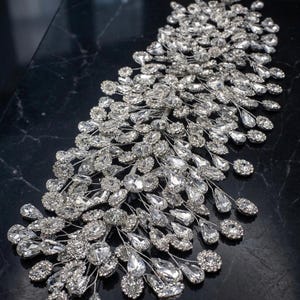 Puede incluir: Un adorno para la cabeza de novia plateado con numerosos cristales transparentes y brillantes. El adorno presenta un diseño asimétrico y fluido con cristales de diferentes tamaños y formas, creando un aspecto glamuroso y elegante. Los cristales están engarzados en delicados alambres plateados.
