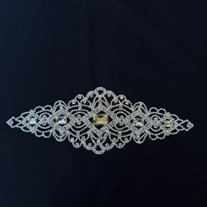 Puede incluir: Una decoración de pedrería plateada con un diseño delicado e intrincado. La decoración presenta una forma de diamante central rodeada de pedrería más pequeña.