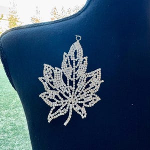 May include: A sparkling, silver-colored rhinestone maple leaf brooch. The brooch is set on a black background. The leaf design is intricately detailed, with each vein and edge outlined with shimmering rhinestones. The brooch has a small loop at the top for attachment.