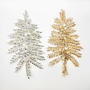 May include: Two decorative Christmas tree ornaments. One is silver with clear crystal accents, and the other is gold with amber crystal accents. Both ornaments are in the shape of a stylized evergreen tree.