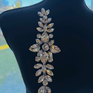 May include: A dazzling crystal rhinestone applique in a vertical design. The applique features clear, faceted rhinestones of various sizes, arranged symmetrically with a central cluster and leaf-like shapes. It's displayed on a black mannequin, showcasing its sparkle.
