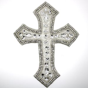Könnte beinhalten: Ein silberfarbenes Kreuz, reich verziert mit klaren, funkelnden Strasssteinen. Das Kreuz hat ein detailliertes Design mit Steinen unterschiedlicher Größe, was einen schillernden Effekt erzeugt. Der Hintergrund ist schlicht weiß.