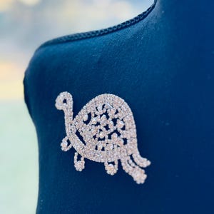 May include: A silver-toned rhinestone turtle brooch. The brooch is attached to a dark blue fabric. The turtle design has a long neck, small legs, and a decorative shell. The brooch is a detailed, sparkling accessory.
