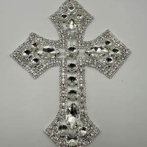 Op de afbeelding: Een decoratief kruis met een sprankelend ontwerp. Het kruis is versierd met talrijke heldere, facetgeslepen strass-steentjes, waardoor een briljant, opvallend effect ontstaat. De kruisvorm is symmetrisch, waarbij elke arm en de top zijn versierd met dezelfde sprankelende stenen.