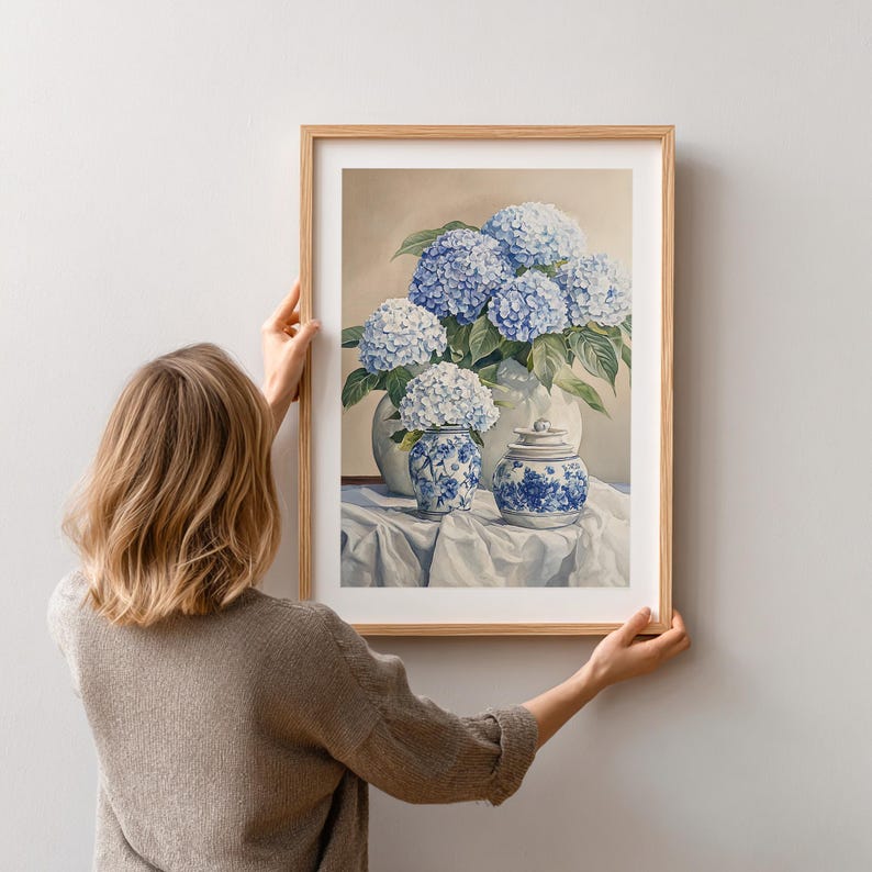 Person hanging framed hydrangea print on wall, showcasing scale and styling of blue floral artwork. French farmhouse decor print with vintage-style hydrangea flower painting.