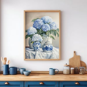 Still life hydrangea artwork in light wood frame styled above kitchen counter with blue cabinets and pottery. Cottagecore decor wall art showcasing detailed blue hydrangea botanical painting.