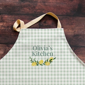 Könnte beinhalten: Hellgrüne und weiße karierte Schürze mit dem Text "Olivia's Kitchen" und einem Zitronen- und Blattdesign. Die Schürze hat ein beiges Nackenband und ist aus Stoff.