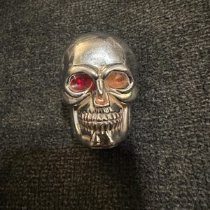May include: A silver skull-shaped object with red and orange eye sockets and a detailed jawline. The skull is set against a dark gray textured background. The object appears to be a decorative item or a piece of jewelry.