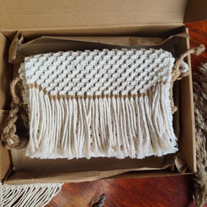 Può includere: Una borsa in macramé bianca con una frangia marrone. La borsa è in una scatola di cartone con carta velina marrone.