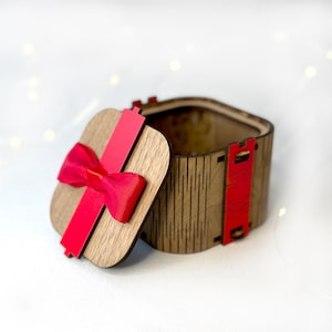 May include: Wooden gift box with a square lid, featuring a red ribbon and bow. The box is made of light brown wood with a decorative pattern. The lid is slightly ajar, revealing the interior.