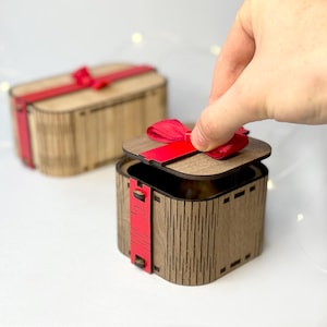 Può includere: Scatole regalo in legno con dettagli di nastro rosso. Una scatola è aperta, rivelando l'interno. Le scatole hanno una finitura a venatura naturale e una forma rettangolare con bordi arrotondati. Il nastro rosso è legato con un fiocco.