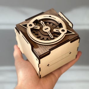 May include: A wooden cube-shaped box with a laser-cut design on the top. The design features a compass-like element with gears and a pointer. The box is held in a hand, showcasing the intricate craftsmanship and detail of the wooden construction.