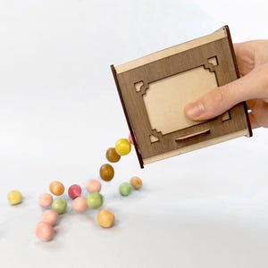 以下が含まれることがあります： 装飾的な切り抜きデザインの木箱が傾いており、カラフルなキャンディーがこぼれています。箱は明るい色の木材でできており、濃い茶色のトリムが付いています。さまざまなパステルカラーのキャンディーが箱の周りに散らばっています。