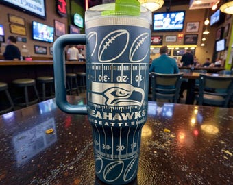 Może przedstawiać: Granatowy kubek termiczny z uchwytem, ​​z motywem boiska do futbolu amerykańskiego i logo Seattle Seahawks. Kubek ma zieloną pokrywkę i stoi na drewnianym stole.