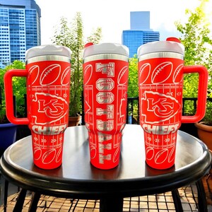 May include: Three red insulated tumblers with white lids and handles. Each tumbler features a football field design with the words "FOOTBALL" and the Kansas City Chiefs logo. The tumblers have measurements in centimetres.