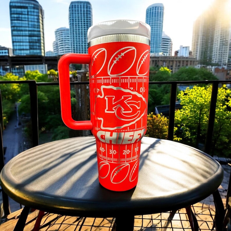 May include: A red insulated tumbler with a large handle, featuring the Kansas City Chiefs logo and football field markings. The tumbler has a white lid and is decorated with silver footballs. The word "CHIEFS" is printed on the tumbler.