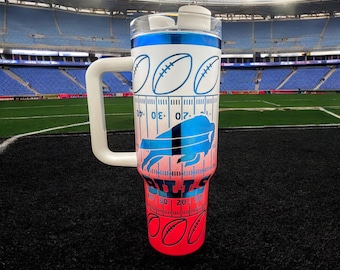 May include: A tall insulated tumbler with a white handle and a clear lid. The exterior features a blue and red gradient design with a football field yard line pattern, football graphics, a blue buffalo logo, and the word BILLS in blue text.