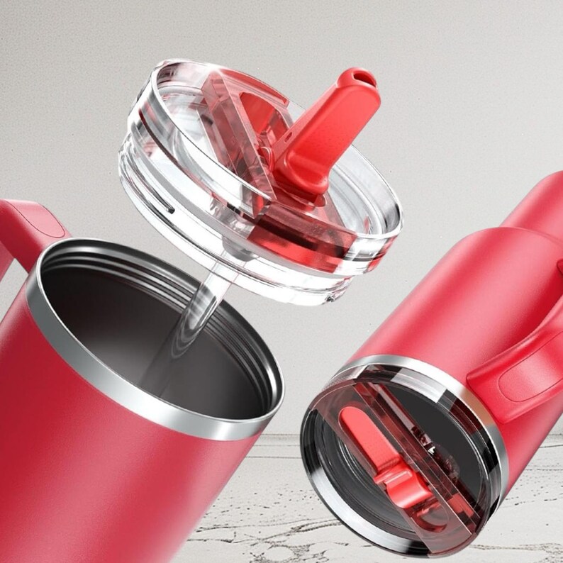 May include: Red insulated tumbler with a clear lid and a red flip-top. The tumbler has a silver rim and a handle. The lid is partially open, revealing the straw. The tumbler is designed to keep beverages cold or hot. The background is blurred.