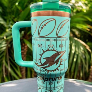 May include: A teal tumbler with a copper lid and a handle. The tumbler has a design of a dolphin, footballs, and a football field with yard lines. The text "Miami Dolphins" is printed on the tumbler.