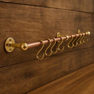 Może przedstawiać: Miedziana i mosiężna szyna kuchenna z ośmioma hakami w kształcie litery S. Szyna jest zamontowana na drewnianej powierzchni. Elementy mosiężne obejmują elementy montażowe i haki, ze środkowym prętem z miedzi.