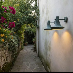 Puede incluir: Dos apliques de pared para exteriores con acabado bronce iluminan un camino de piedra. Las luces están montadas en una pared blanca, y el camino está bordeado de exuberante vegetación y flores coloridas, creando un ambiente cálido y acogedor.