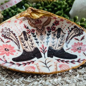 Könnte beinhalten: Eine weiße Keramikschale mit goldfarbenem Rand, verziert mit einem rosafarbenen Blumenmuster und zwei schwarzen Cowboystiefeln.