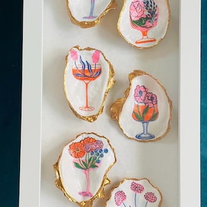 Puede incluir: Un conjunto de seis conchas de ostra, cada una con un borde dorado y un diseño floral único dentro de una copa de cóctel. Los diseños presentan colores vibrantes como el rosa, el naranja y el azul, sobre un fondo blanco.